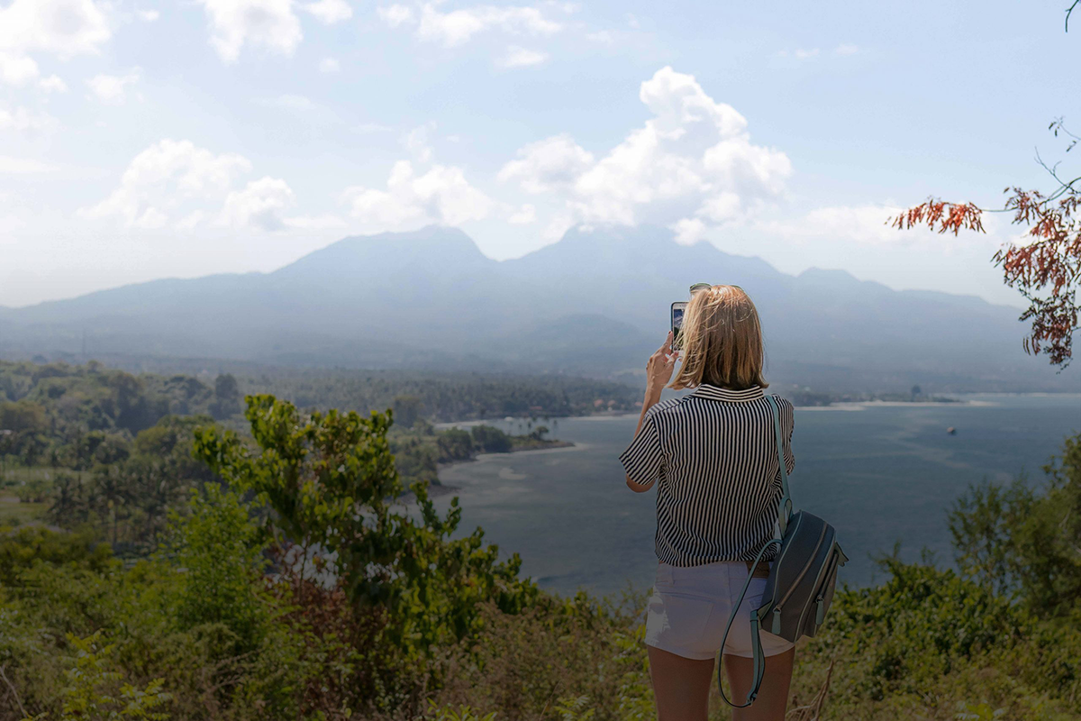 Traveler taking photo on phone of mountains in distance next to ocean