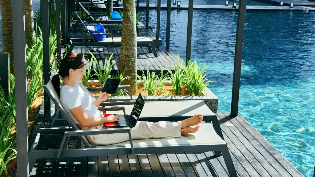 Digital nomad sits on lounge chair next to pool working remotely from laptop and tablet