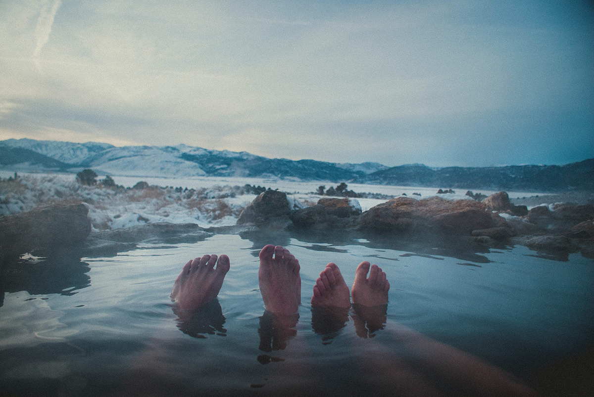 two persons four feet sticking out of a hot spring pool with mountains in background